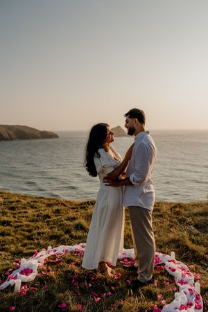 Couple embracing at sunset on coastal cliffs with flower petals arranged on grass and ocean backdrop.