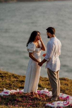 Woman covers mouth in surprise during romantic seaside proposal with rose petals at golden hour.