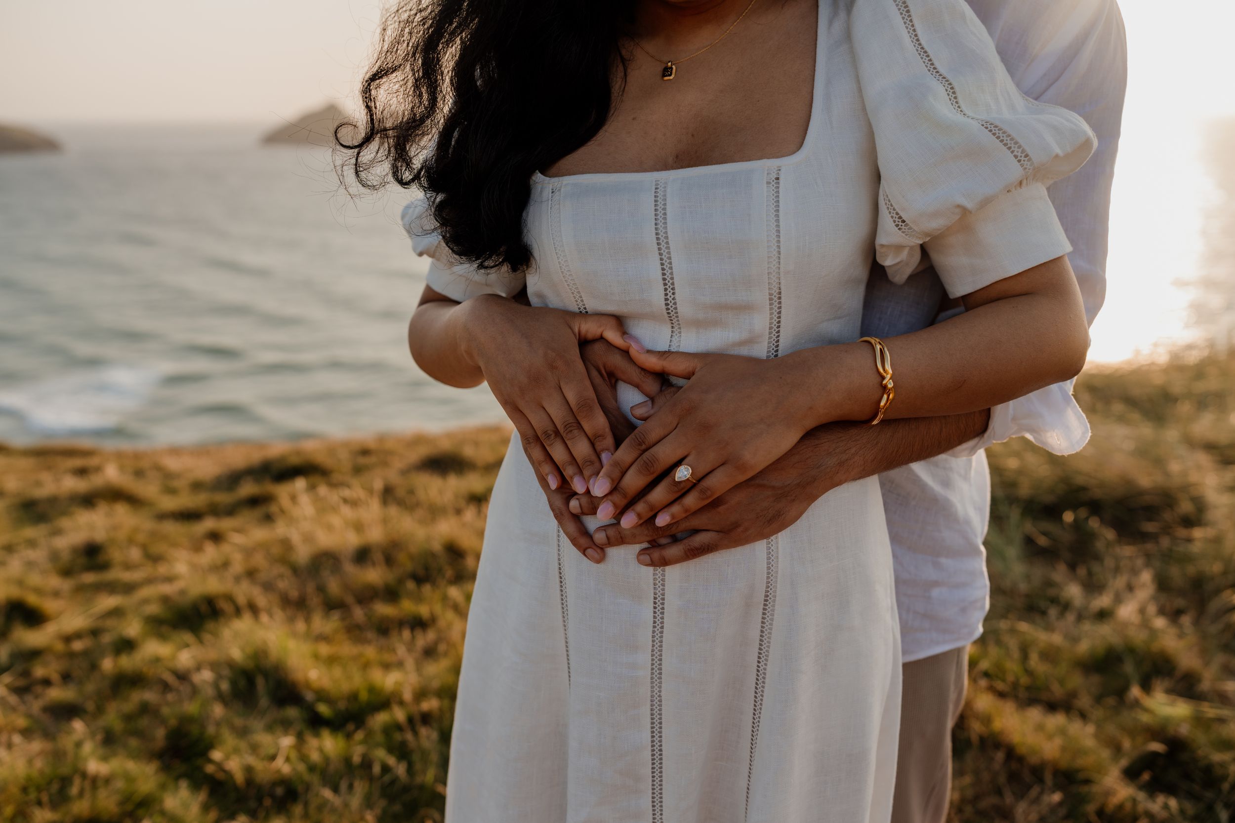 Close-up of couple embracing outdoors, woman in white dress with gold bracelet, ocean backdrop at sunset.