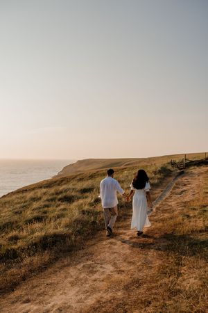 Couple in white outfits holding hands walking along a coastal cliff path at golden hour sunset.