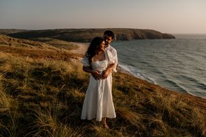 Couple embracing on coastal cliffs at golden hour, woman in white dress, ocean and headland backdrop.