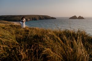 Couple standing on coastal cliffs at golden hour with ocean and headlands in background.