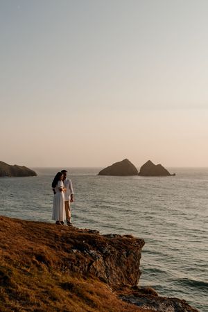 Couple in white standing on rocky coastal cliff at sunset with ocean and rock formations behind them.