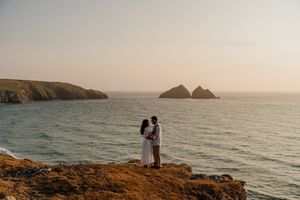 Couple embracing on rocky coastal cliffs at sunset with ocean and islands in background.