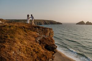 Couple in white standing on a dramatic coastal cliff edge overlooking turquoise ocean waters at golden hour.