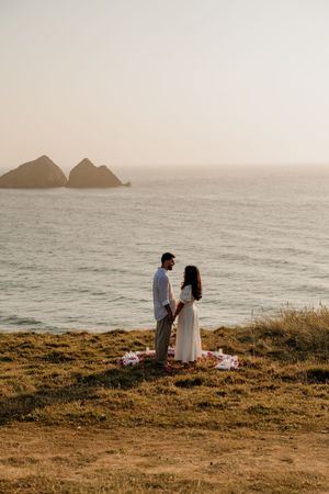 Couple holding hands on coastal cliffside at sunset with rocky ocean backdrop and picnic blanket.