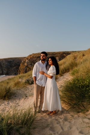 Couple in white outfits embracing on sandy beach dunes at golden hour with coastal cliffs in background.