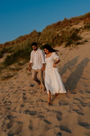 Couple in white outfits holding hands while walking on sandy beach dunes at golden hour.