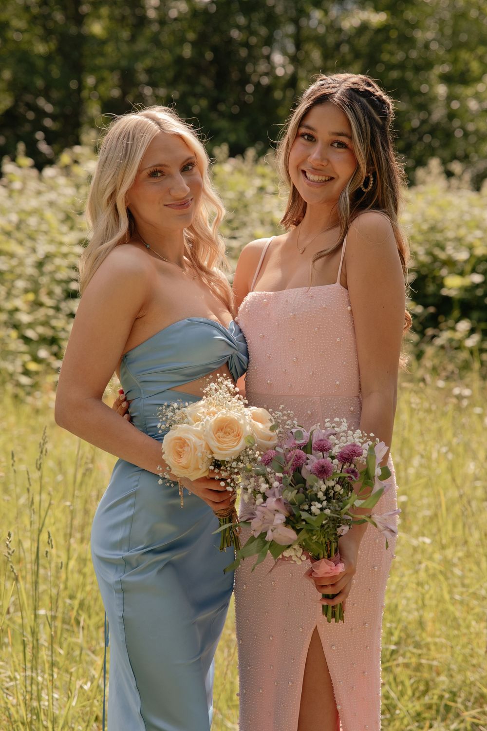 Two bridesmaids in blue and pink gowns holding flower bouquets, posing outdoors in a lush green field.