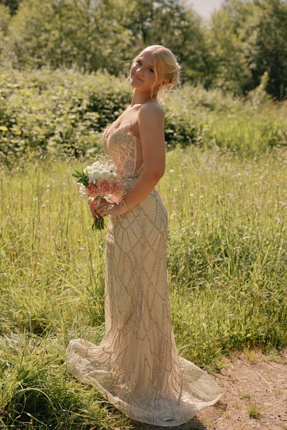 Blonde bride in beaded champagne gown holding white bouquet, smiling outdoors in sunlit green meadow.