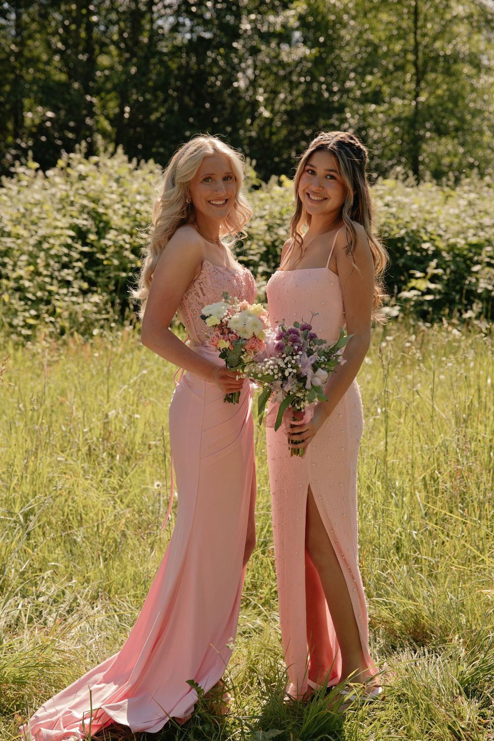 Two bridesmaids in pink satin gowns holding floral bouquets stand in a sunlit meadow surrounded by lush greenery.