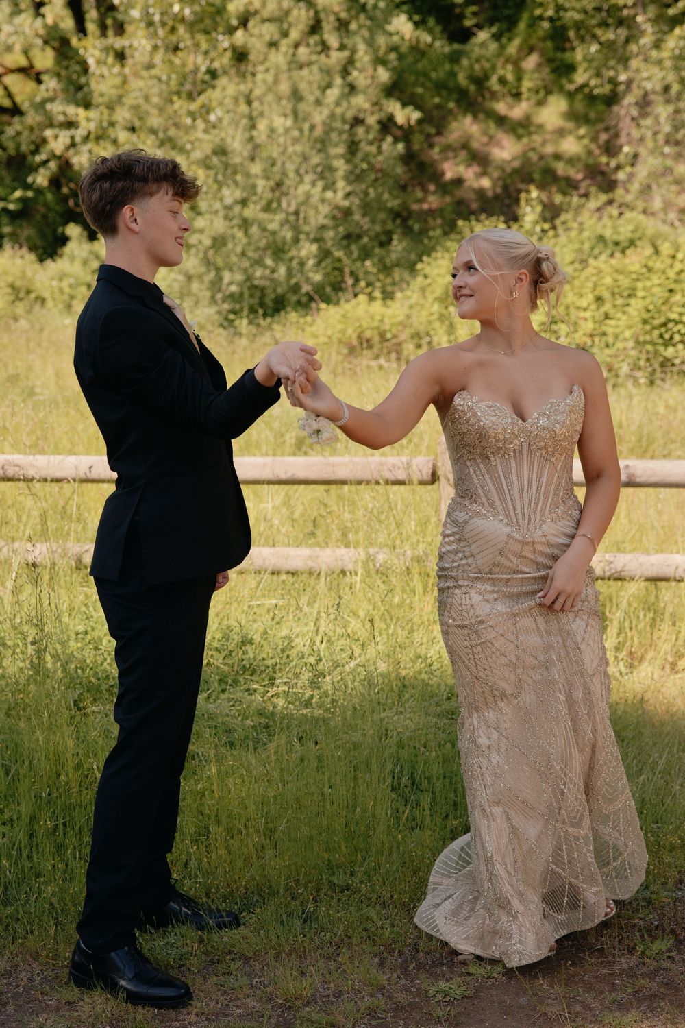 A young couple in formal attire dance outdoors, man in black suit, woman in gold gown, surrounded by lush greenery.