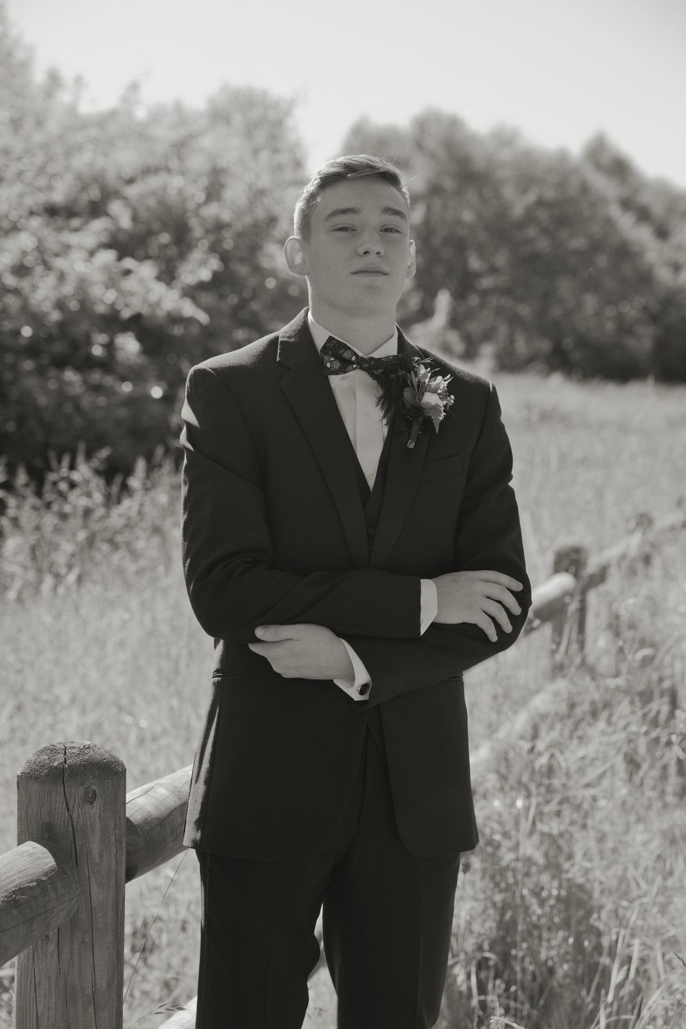 Young man in black suit with floral boutonniere and bow tie poses arms crossed by rustic fence in black and white photo.