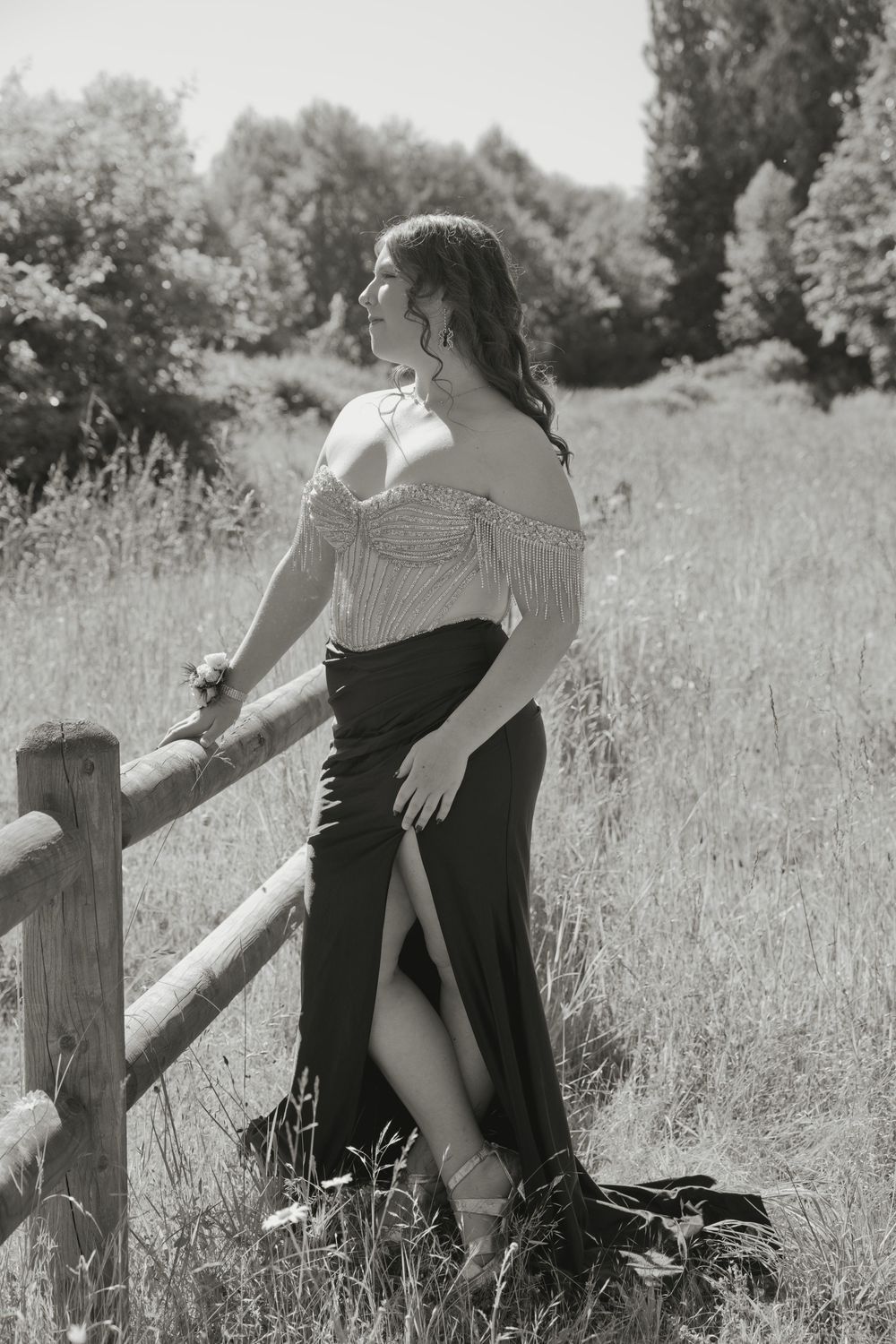 Black and white photo of a woman in an off-shoulder top and slit skirt leaning on a wooden fence in a field.