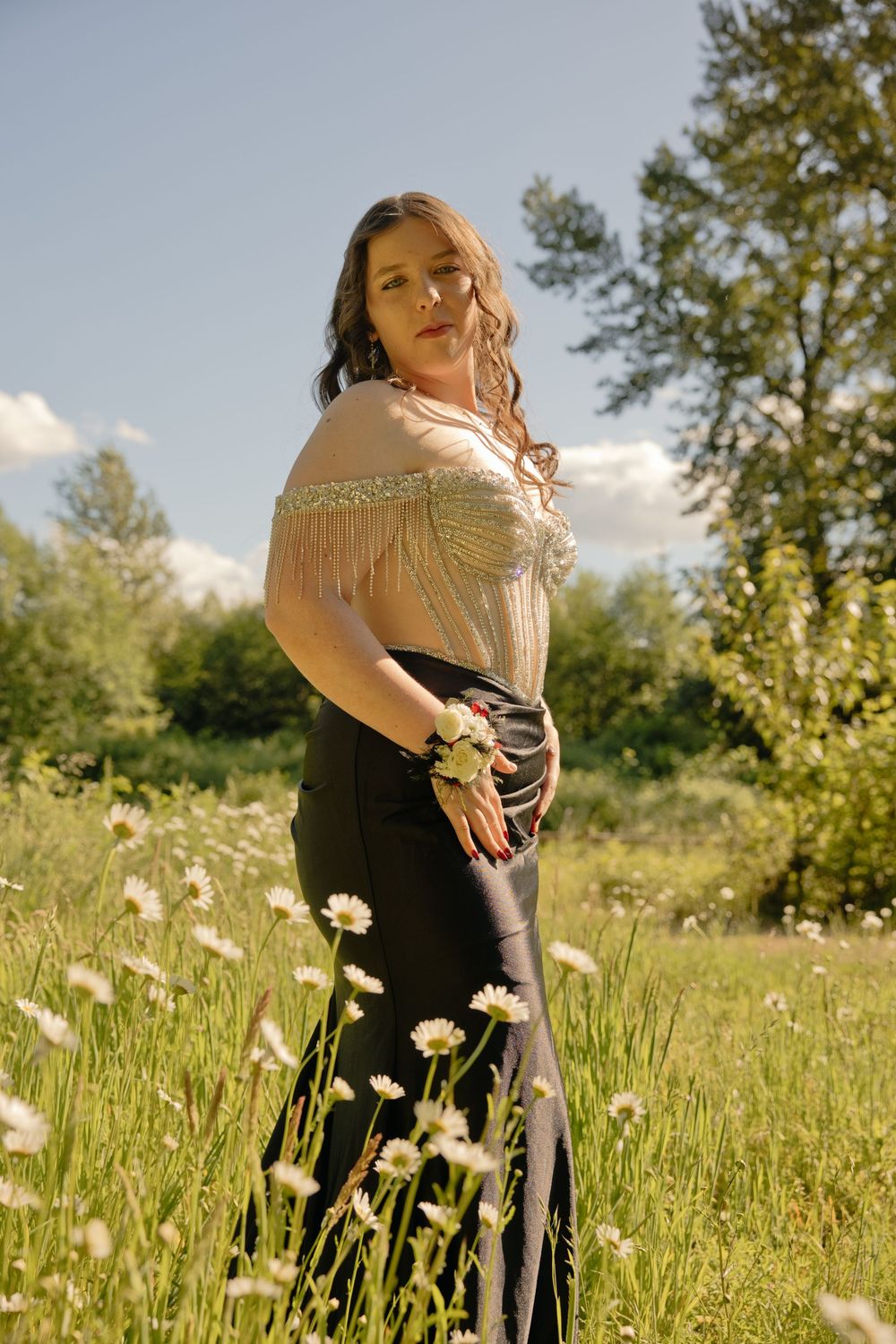 Woman in fringed top and dark skirt stands among white daisies in a sunny green meadow with trees in the background.