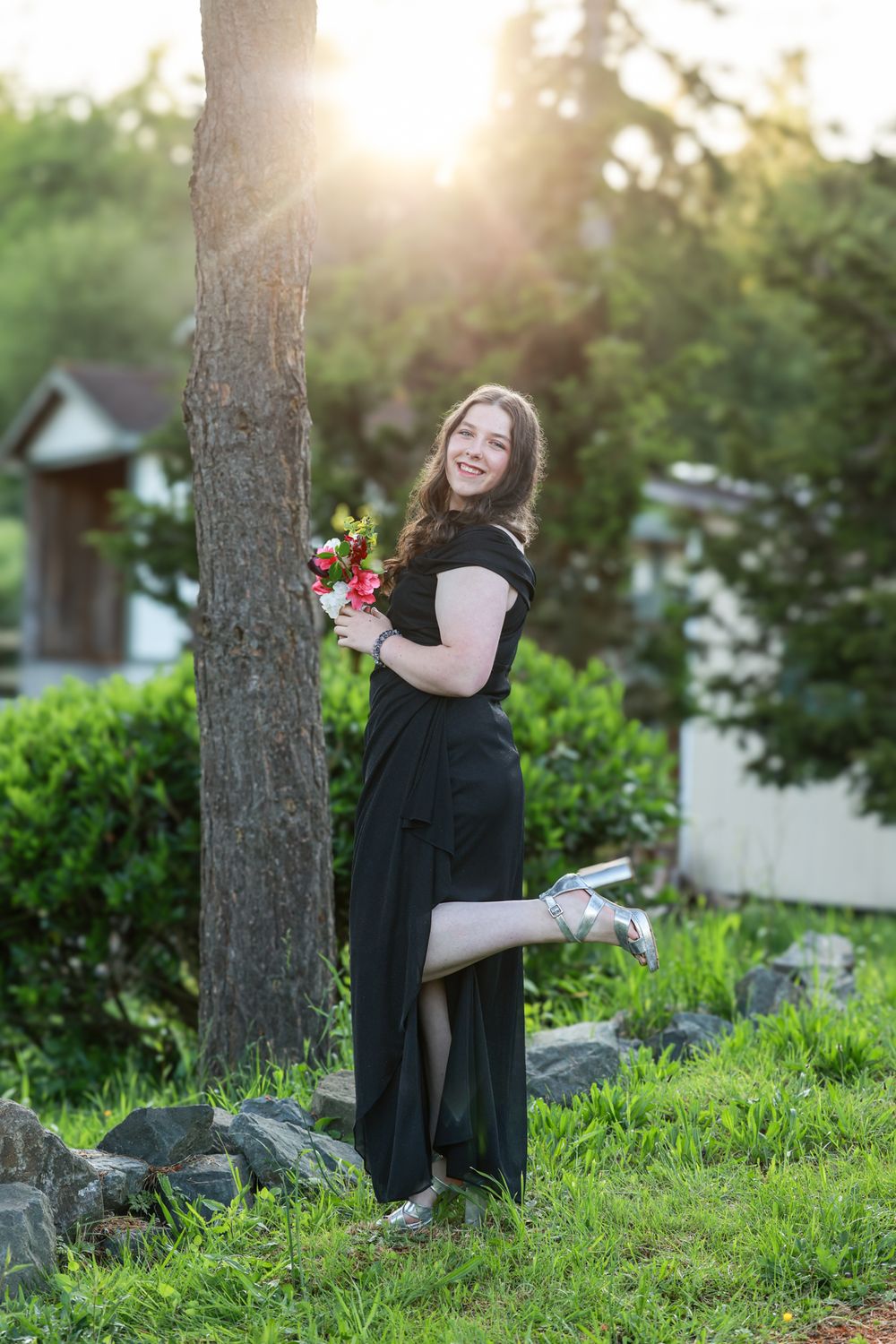 Smiling young woman in black dress holding red bouquet, posing by tree at golden hour sunset in garden.