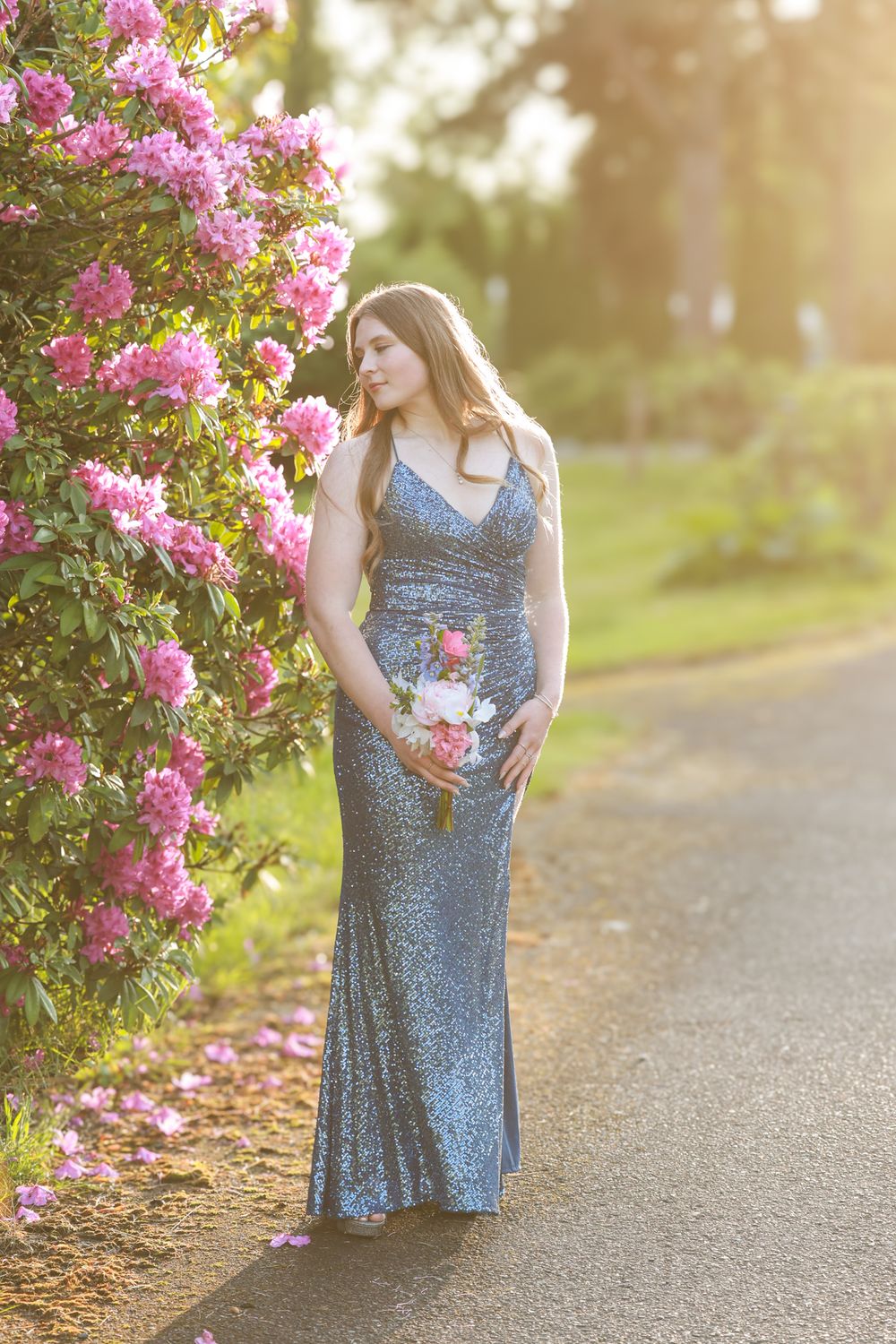 Woman in silver sequin gown holding bouquet beside pink flowering bush in golden sunlight.