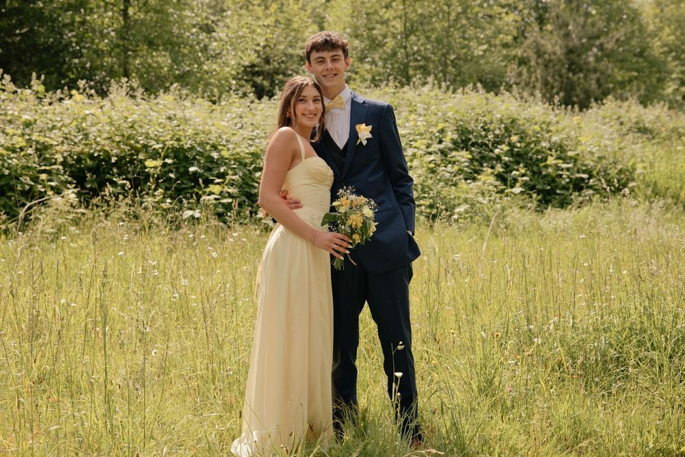 Couple posing in a lush green meadow, woman in yellow gown holding bouquet, man in navy suit, outdoor wedding photo.
