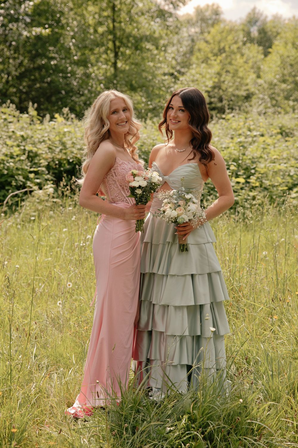 Two women in elegant pink and sage green gowns holding bouquets stand in a lush green meadow at a wedding.
