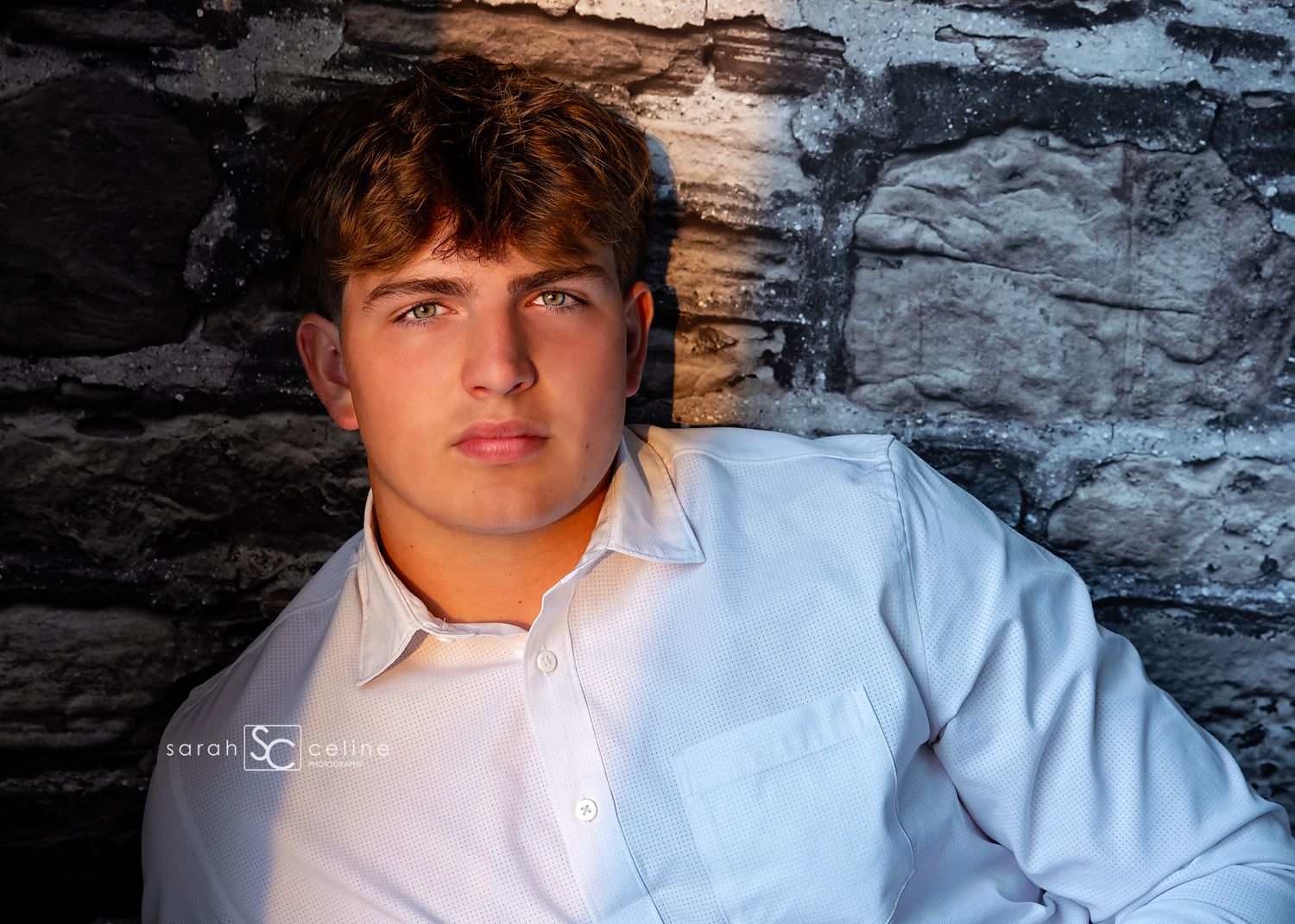 Young man in white shirt poses against stone wall in professional senior portrait photography.