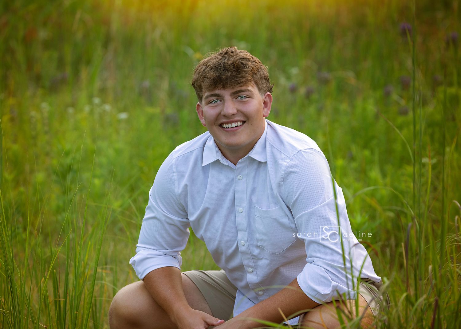 Smiling teenage boy in white shirt sitting in a lush green meadow with wildflowers for senior photos.
