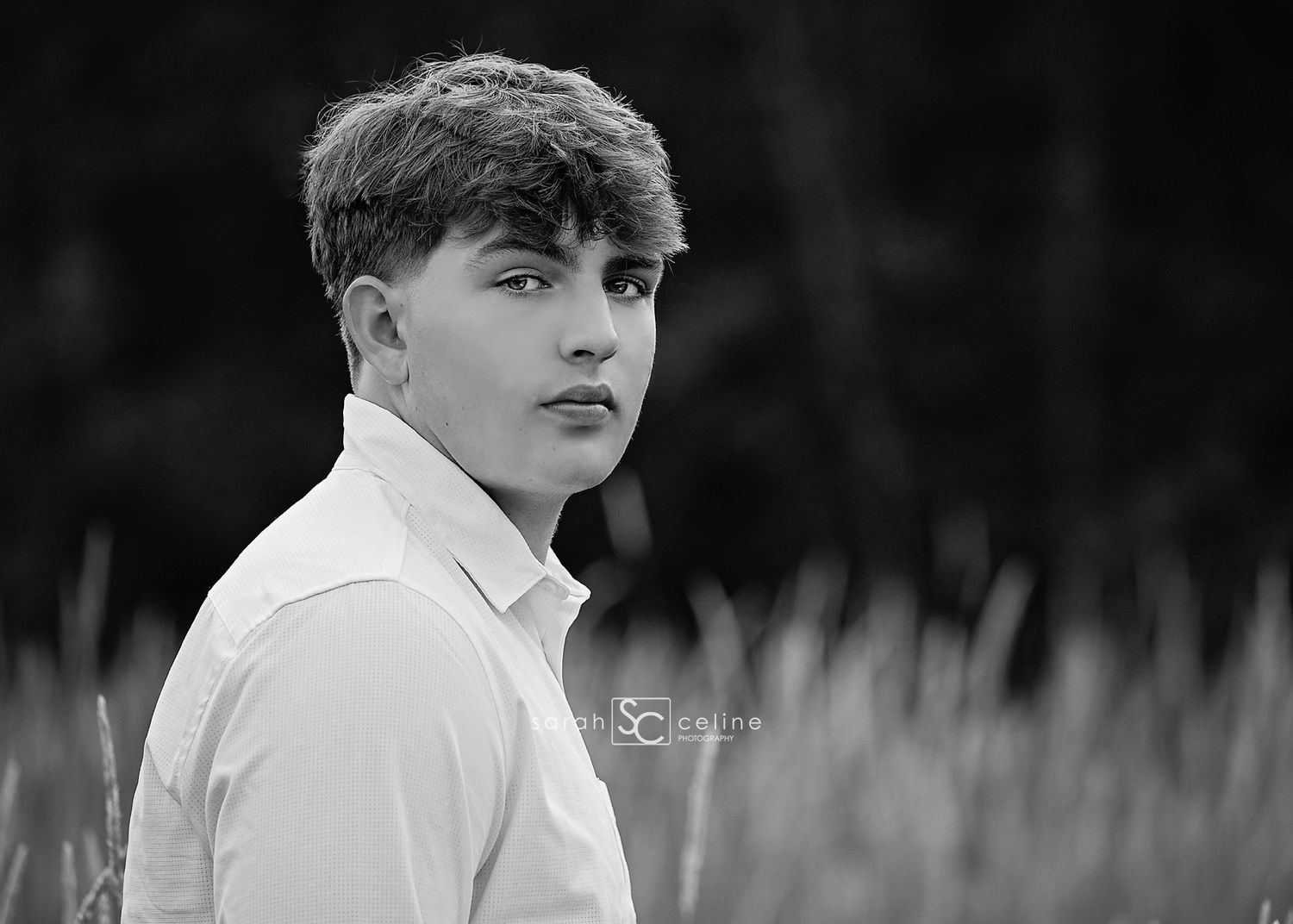 Black and white portrait of a young man in a white shirt gazing thoughtfully, with blurred natural foliage background.