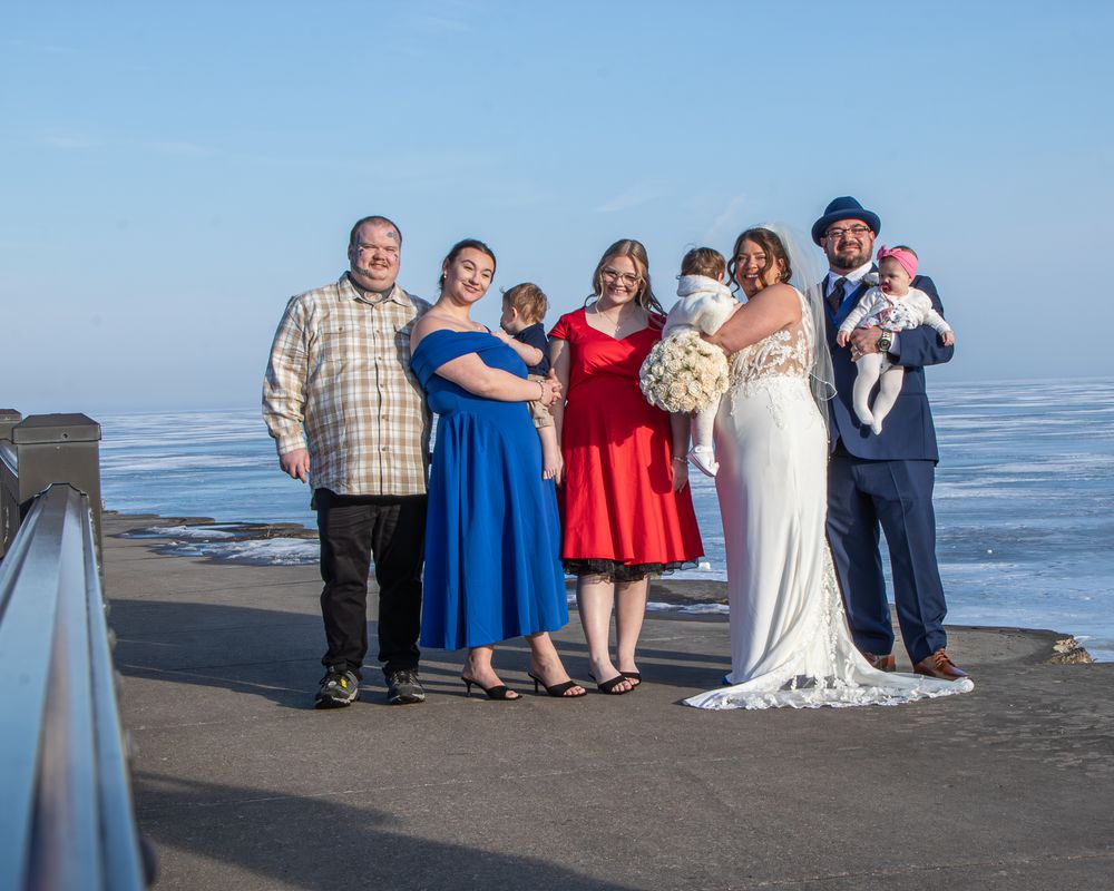 Wedding party poses outdoors by frozen waterfront, bride in white gown with bouquet, guests in blue, red attire.