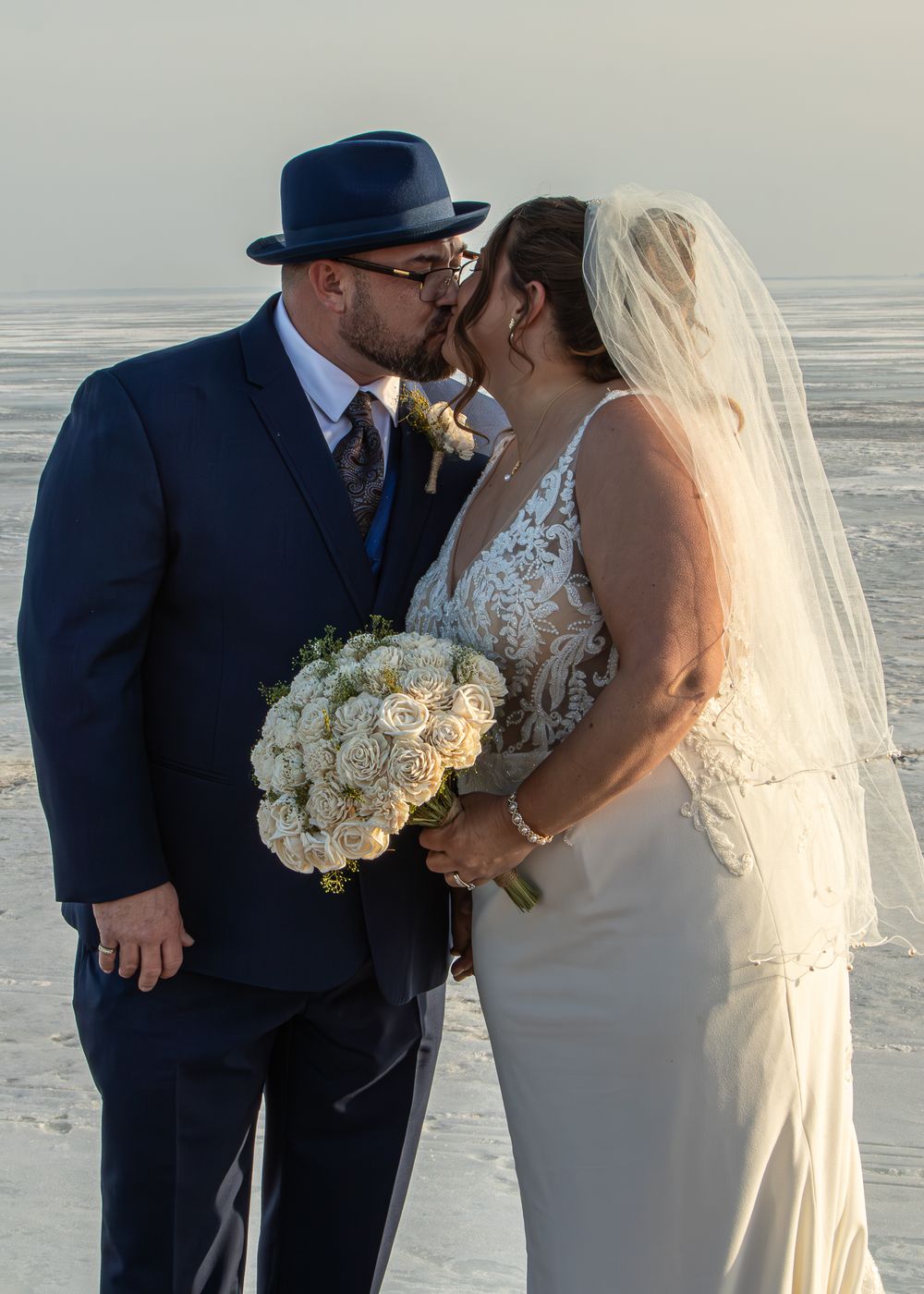 Bride and groom share a kiss on the beach, she holds white roses bouquet, he wears navy suit and hat.