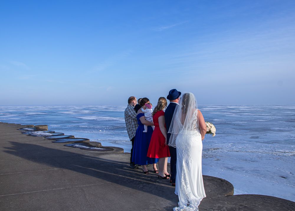 Small wedding ceremony on a frozen lakeside path, bride in white gown holding bouquet with guests in colorful attire.