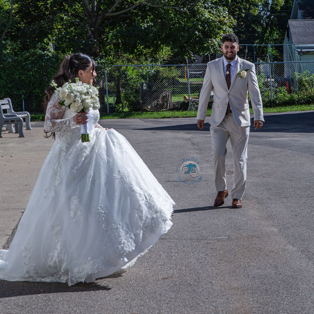 A bride in a white ball gown holding a bouquet walks with a groom in a gray suit outdoors on a sunny day.