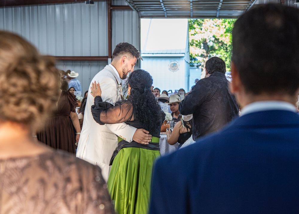Couple sharing an emotional hug during a wedding reception in a rustic venue with guests watching.