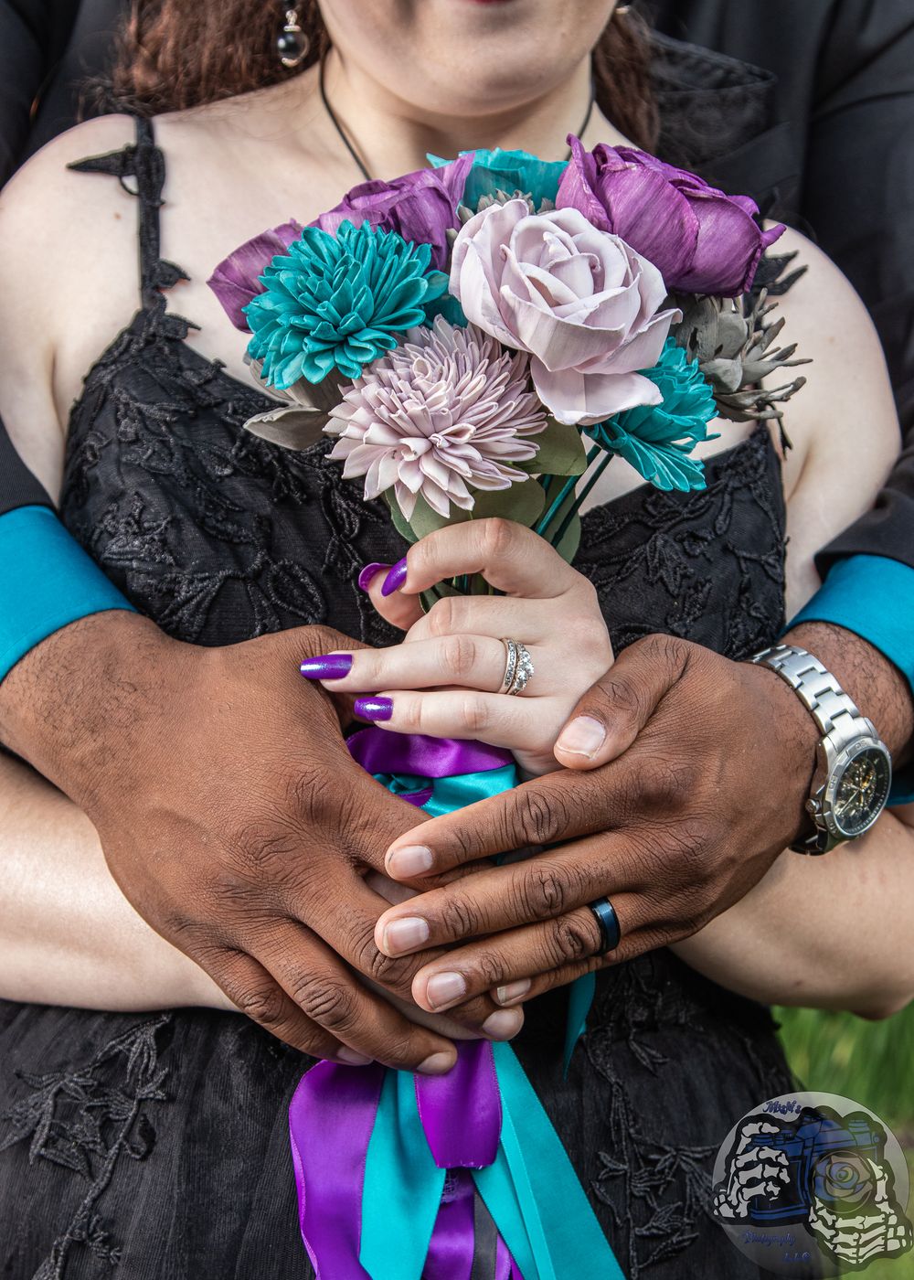 Interracial couple holding teal and purple wedding bouquet, wearing rings, bride in black lace dress.