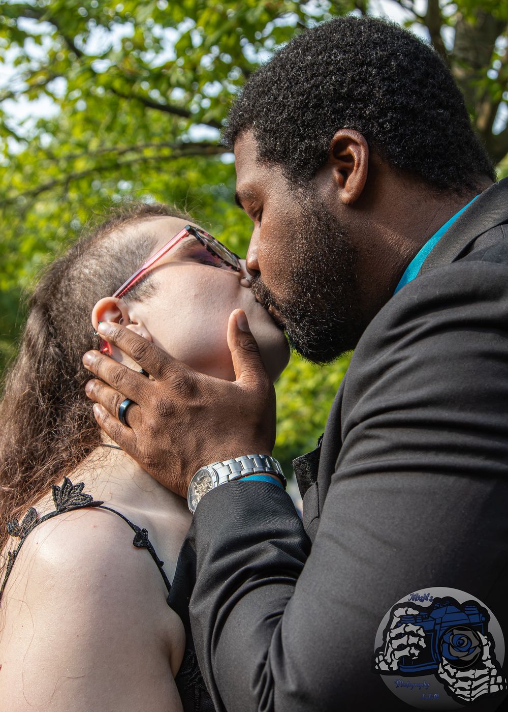 A couple shares a romantic kiss outdoors, with the man in a dark suit gently holding the woman's face.