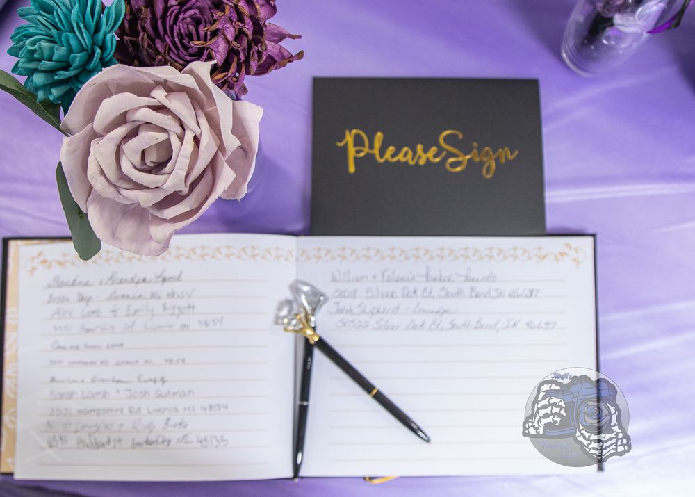 Wedding guest book open on purple table with "Please Sign" card, black pen, and paper flower bouquet.