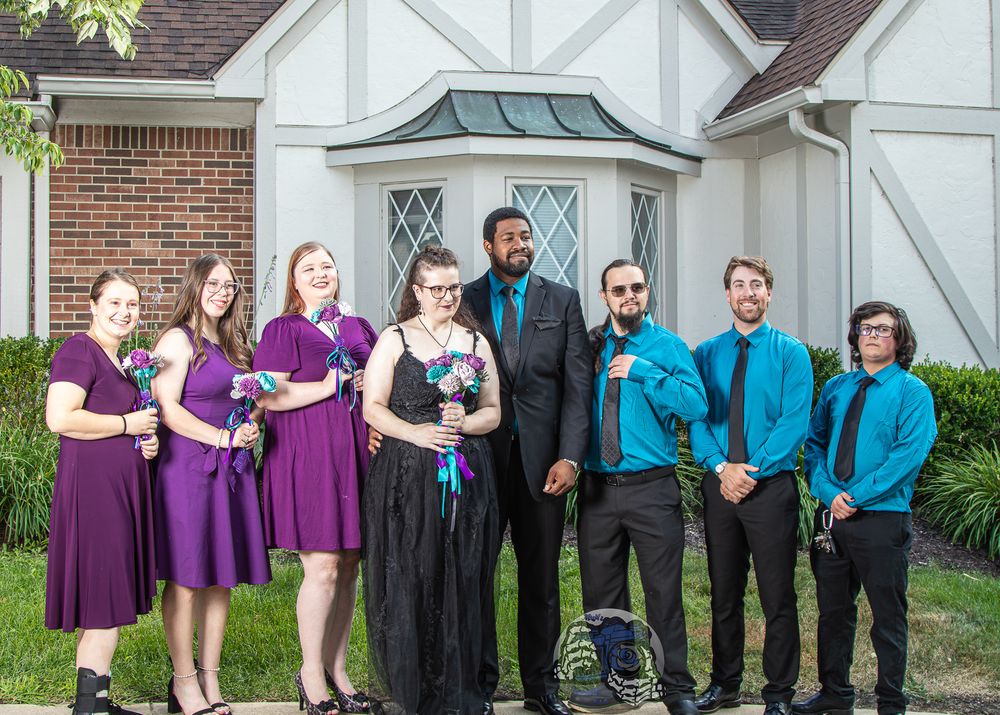 Wedding party posing outside church, bridesmaids in purple dresses, groomsmen in teal shirts and black pants.