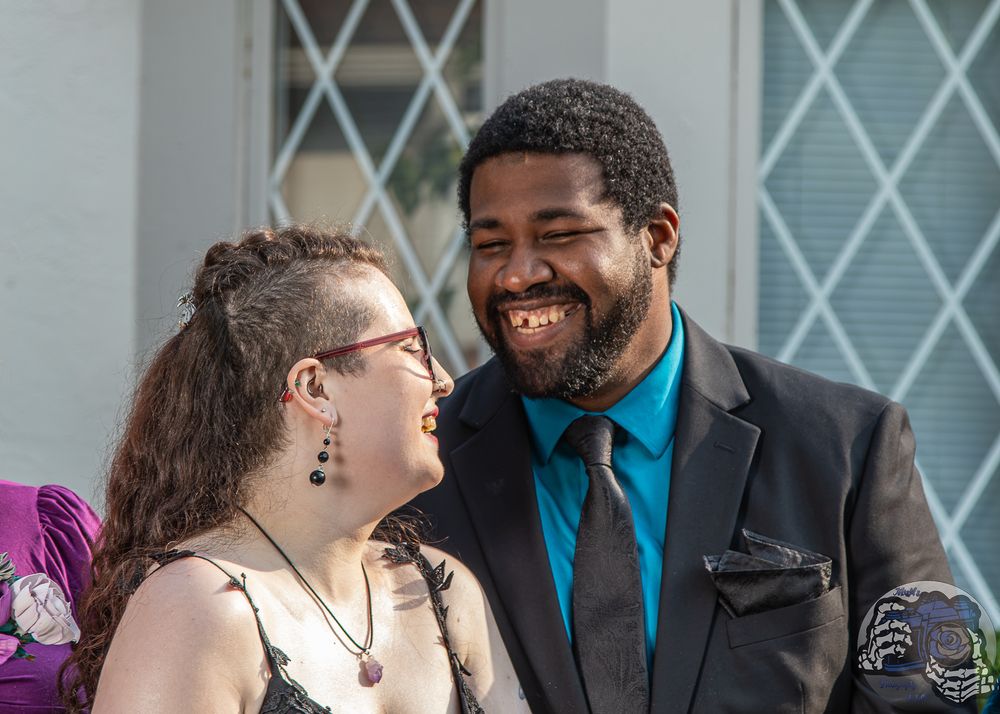 A joyful couple laughing together outdoors, man in teal shirt and blazer, woman in spaghetti strap top with earrings.