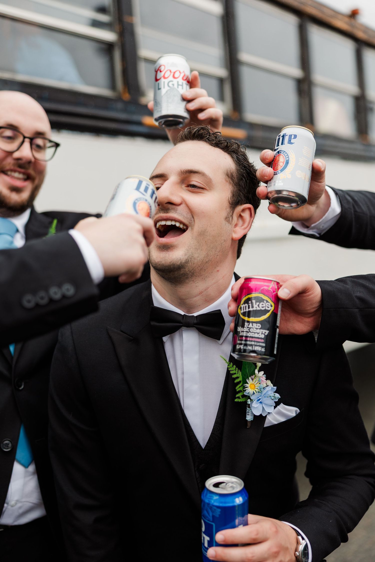 Groom in tuxedo surrounded by groomsmen holding beer cans at wedding celebration outdoors.