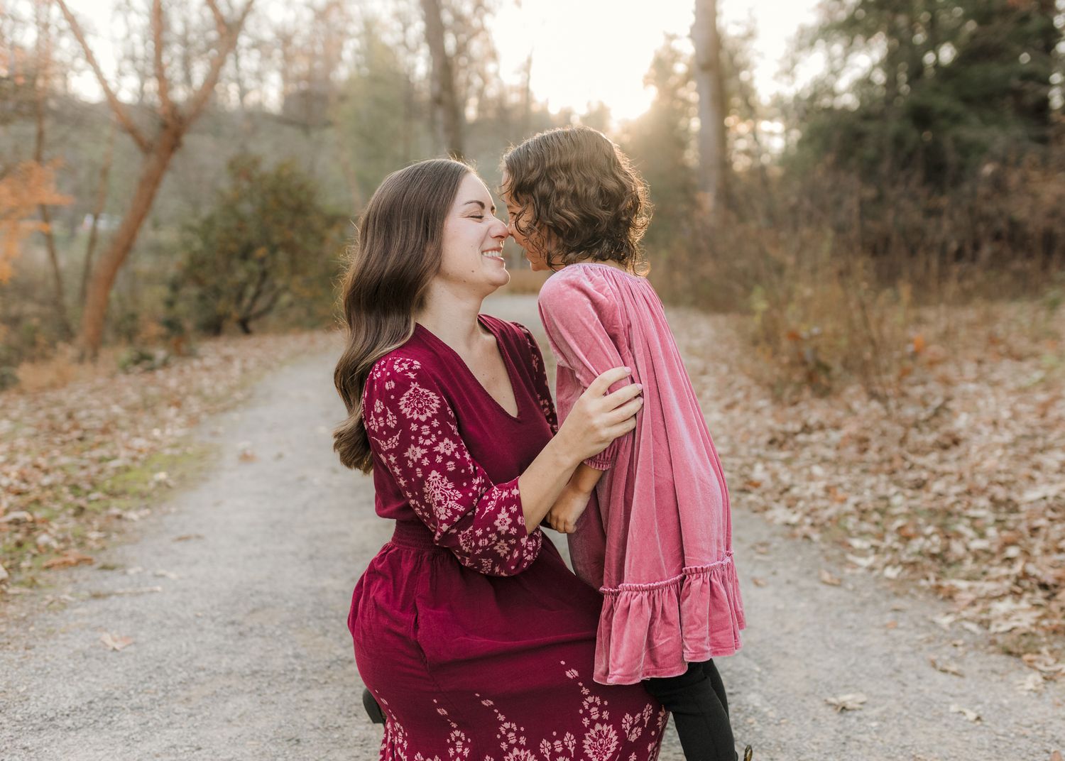 A joyful mother and daughter pose outdoors, with a laughing woman in a red dress nose tickles a girl in a pink dress during golden hour.