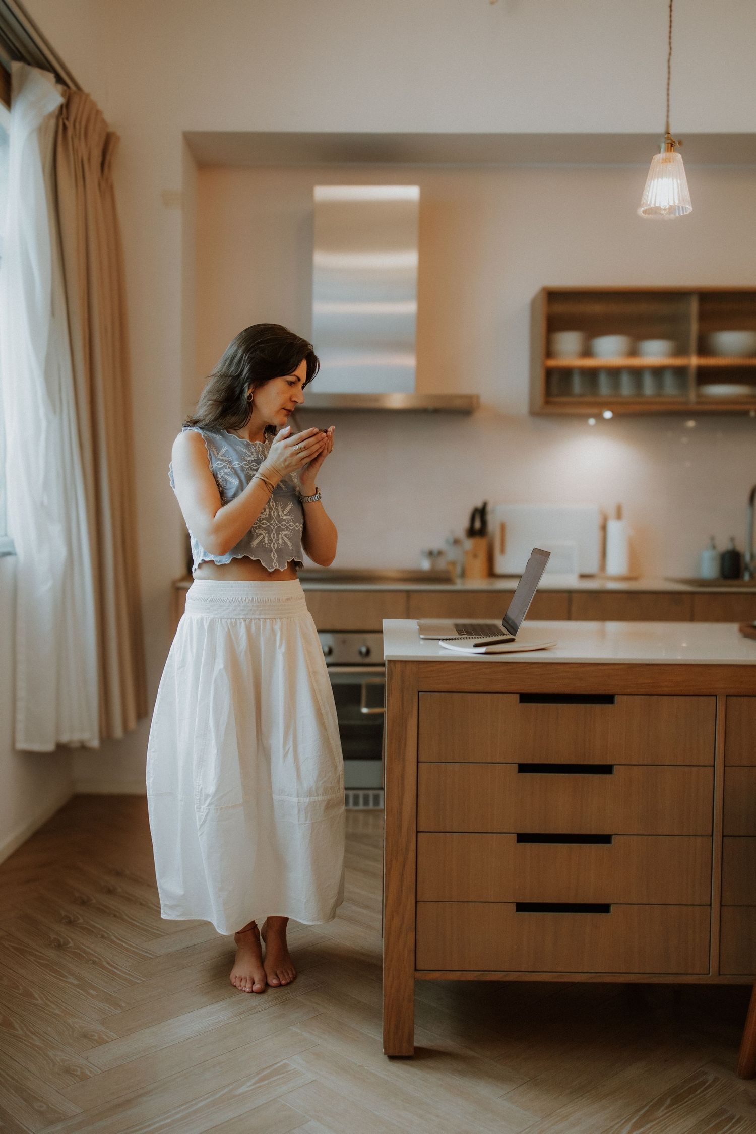 Woman in white skirt sipping coffee while working on laptop at wooden kitchen island in modern home.