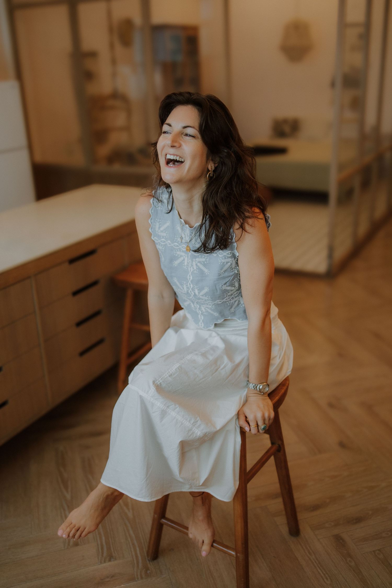 Woman in white skirt and blue top laughing joyfully while seated on a wooden stool in a modern interior.