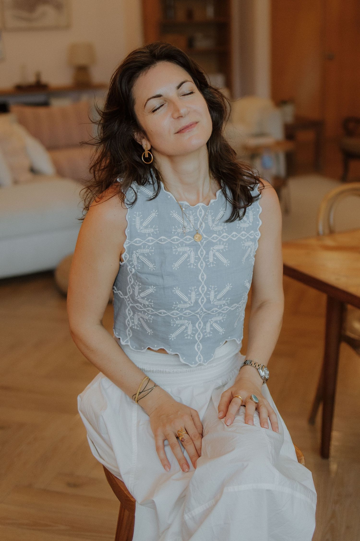 Woman in blue embroidered top and white skirt sits peacefully with eyes closed in a warmly lit living room.