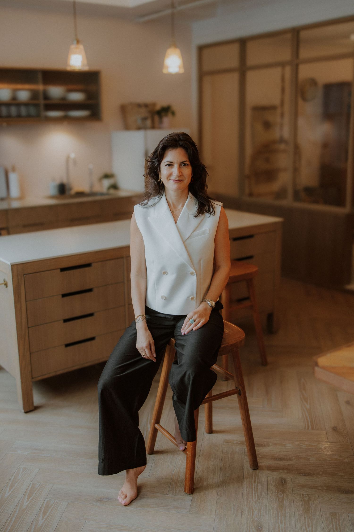 Woman in white blazer and black pants sits on wooden stool in modern kitchen with warm pendant lighting.