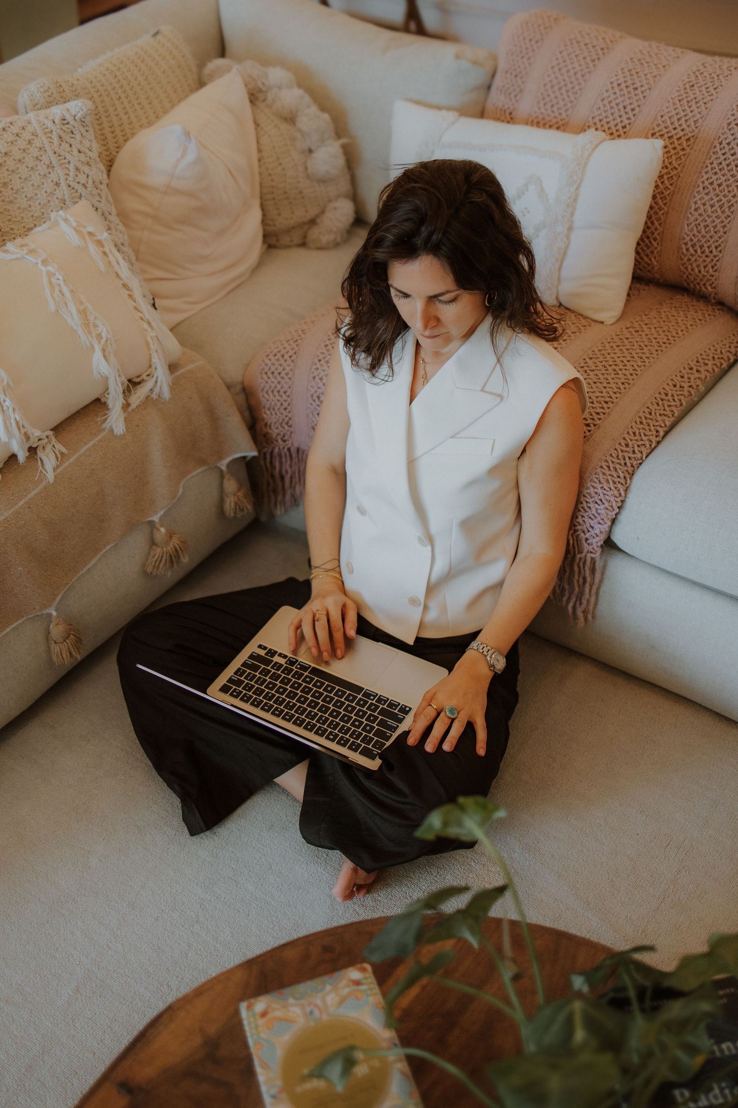 Woman sitting cross-legged on floor using laptop, leaning against white sofa with boho cushions in cozy living room.