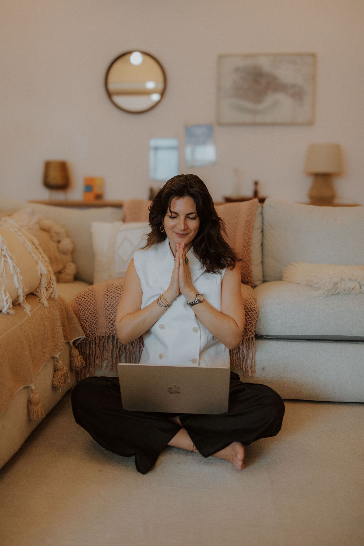 Woman sitting cross-legged on floor with laptop, hands in prayer pose, in cozy boho living room.