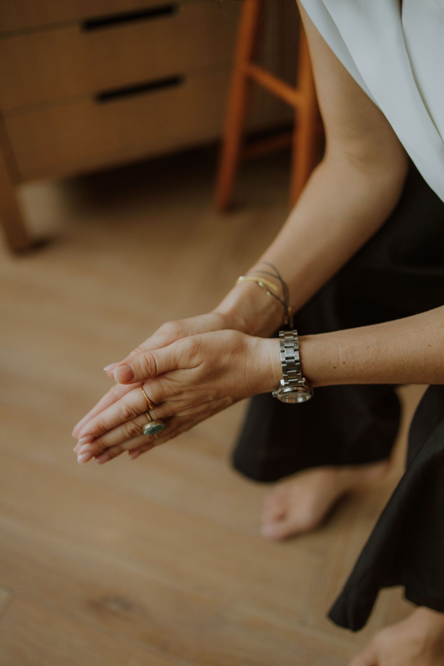 Close-up of woman's hands wearing a silver watch, gold bracelet, and rings, seated on hardwood floor.