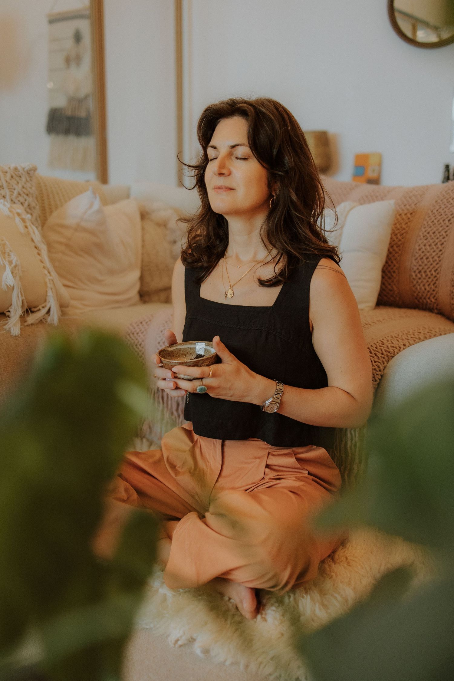 Woman meditating cross-legged on floor, eyes closed, holding singing bowl, in cozy boho living room.