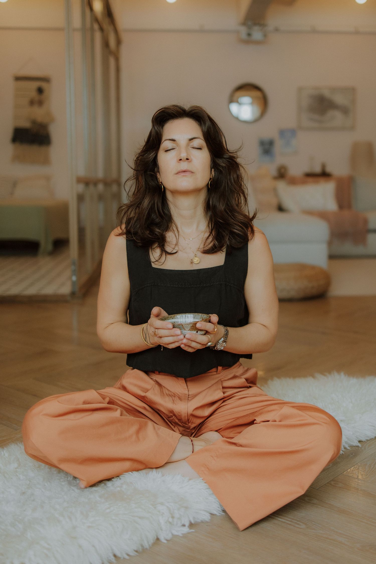 Woman meditating in lotus pose on white rug, holding singing bowl, wearing orange pants and black top.