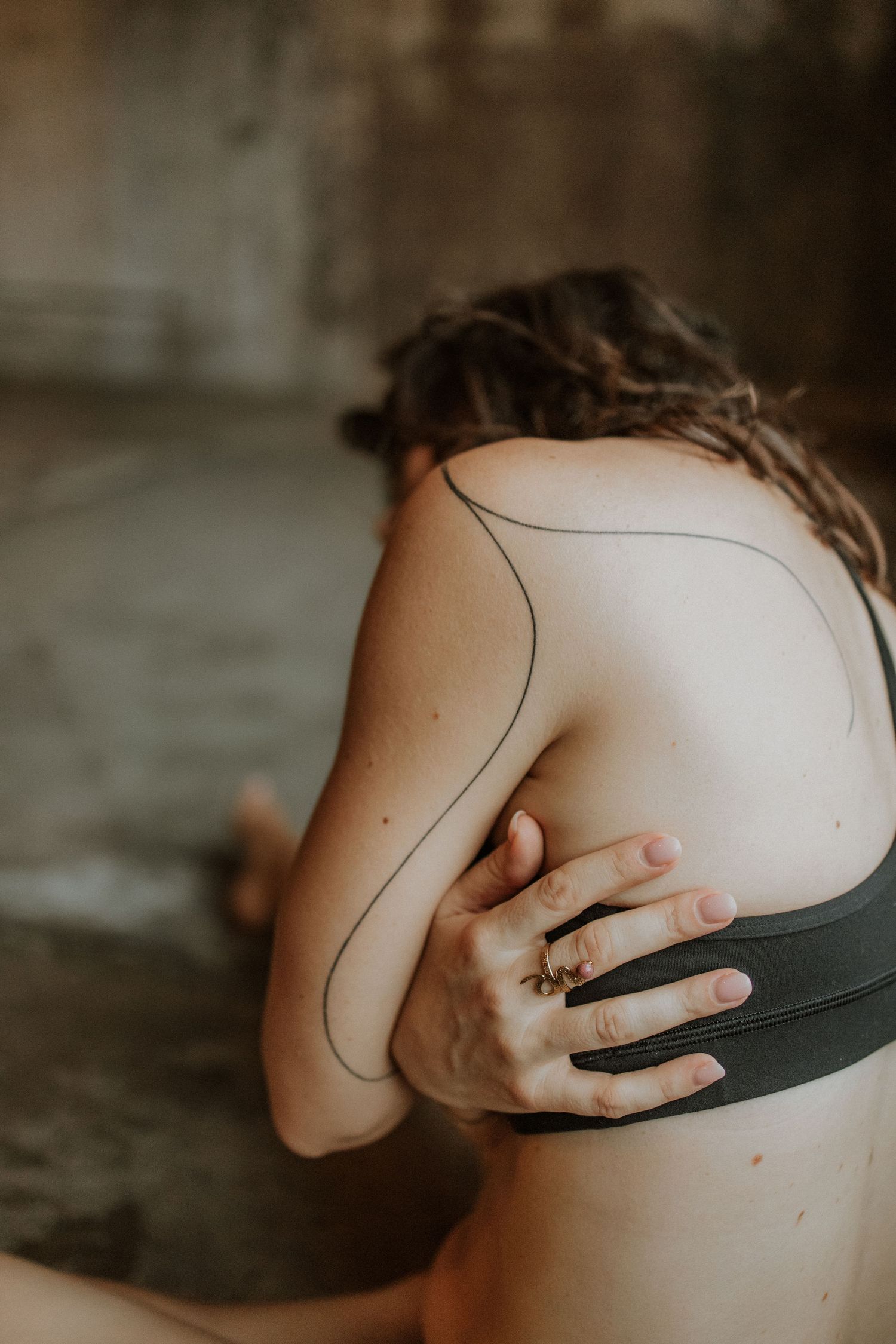 Person with a minimalist line tattoo on their back, wearing a black sports bra, with hand resting on shoulder.