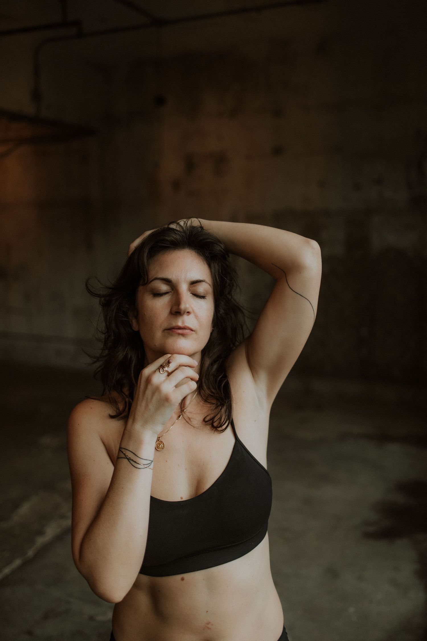 Woman in black crop top poses with eyes closed and hand in hair in a moody, dimly lit industrial space.