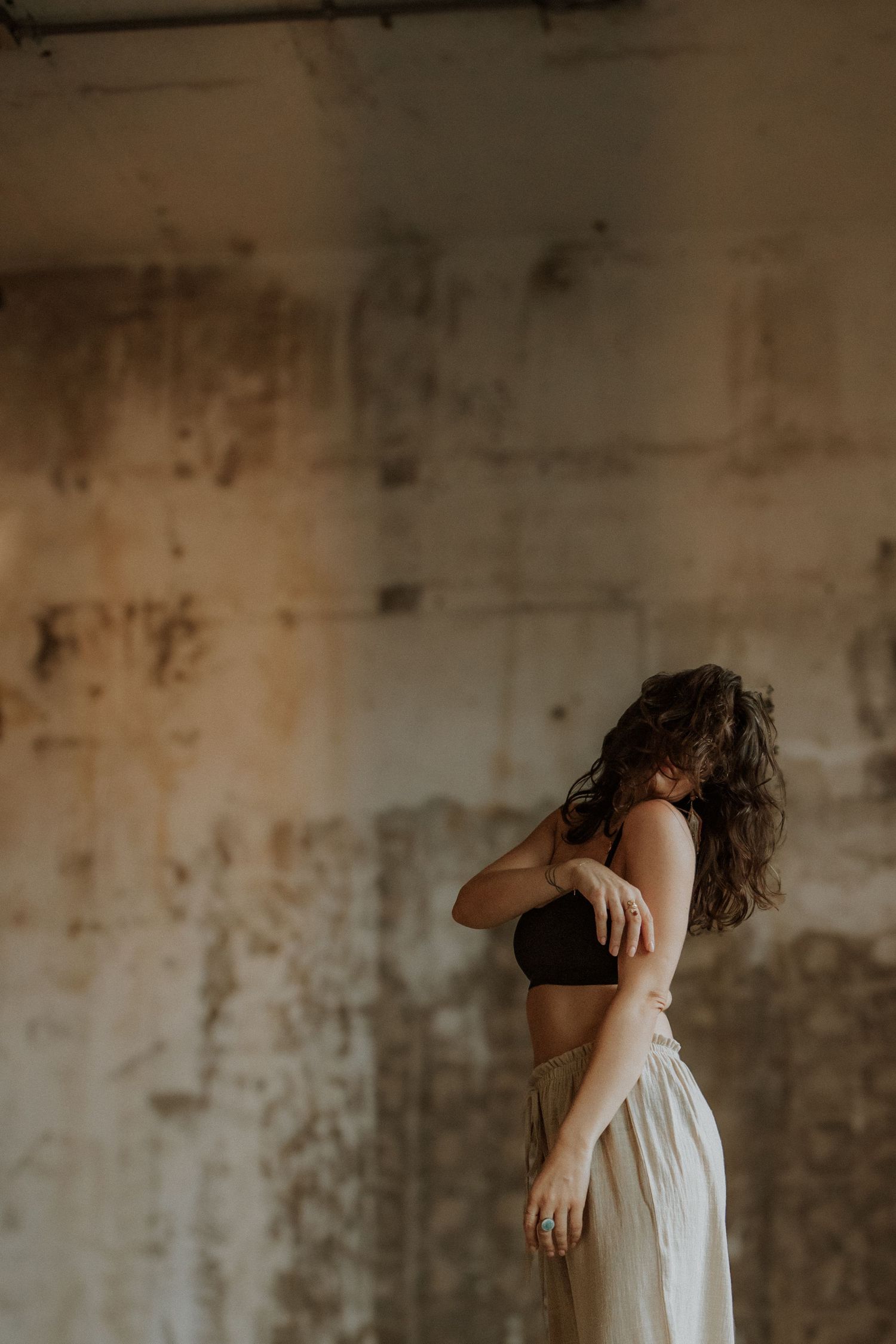 Woman with curly hair in black crop top and flowing cream skirt poses gracefully against a rustic concrete wall.